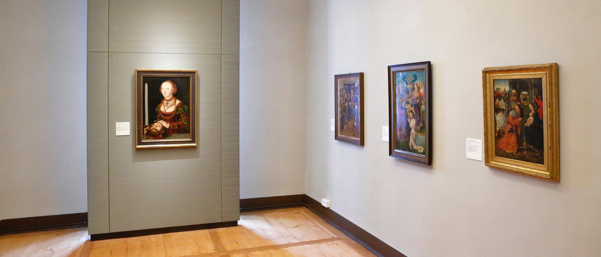 Blick in einen Kunstausstellungsraum. An der rechten Wand hängen drei gotische Tafelgemälde. An der Wand links davon hängt das Gemälde Judith mit dem Haupt des Holofernes von Lucas Cranach dem Älteren.