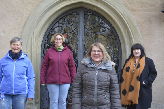 Gruppenbild der vier Museumspädagoginnen. Alle vier lächeln in die Kamera. Links steht Renate Kiesel. Sie trägt eine blaue Jeanshose, eine blaue Jacke und einen dunklen Schal. Sie hat eine graue Kurzhaarfrisur. Rechts neben ihr steht Beatrice Rose-Ebel. Sie trägt eine hellblaue Jeanshose, eine dunkelrote Jacke und einen hellgrünen Schal. Sie hat dunkelblonde Haare, die bis zu den Ohren reichen. Susanne Bocklet steht rechts neben ihr. Sie trägt eine dunkelgraue Jacke. Ihre halblangen, dunkelblonden Haare trägt sie offen. Ganz rechts steht Julia Böhler. Sie trägt einen schwarzen Mantel und einen großen braunen Schal mit großen schwarzen Punkten. Ihre halblangen, schwarzen Haare trägt sie offen.