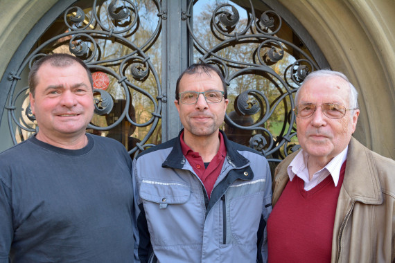 Gruppenbild mit drei Männern. Links im Bild steht der Gärtner Andreas Then. Er trägt ein schwarzes T-Shirt und eine Kurzhaarfrisur. In der Mitte steht der Hausmeister Wolfgang Lochner. Er trägt ein dunkelrotes Polo-Shirt und darüber eine blau-schwarze Arbeitsjacke. Er hat eine Brille und dunkle, kurze Haare. Rechts steht Günter Falkenberg. Er trägt ein weißes Hemd, einen dunkelroten Pullover und eine hellbraune Jacke. Er hat eine Brille und graue, kurze Haare.