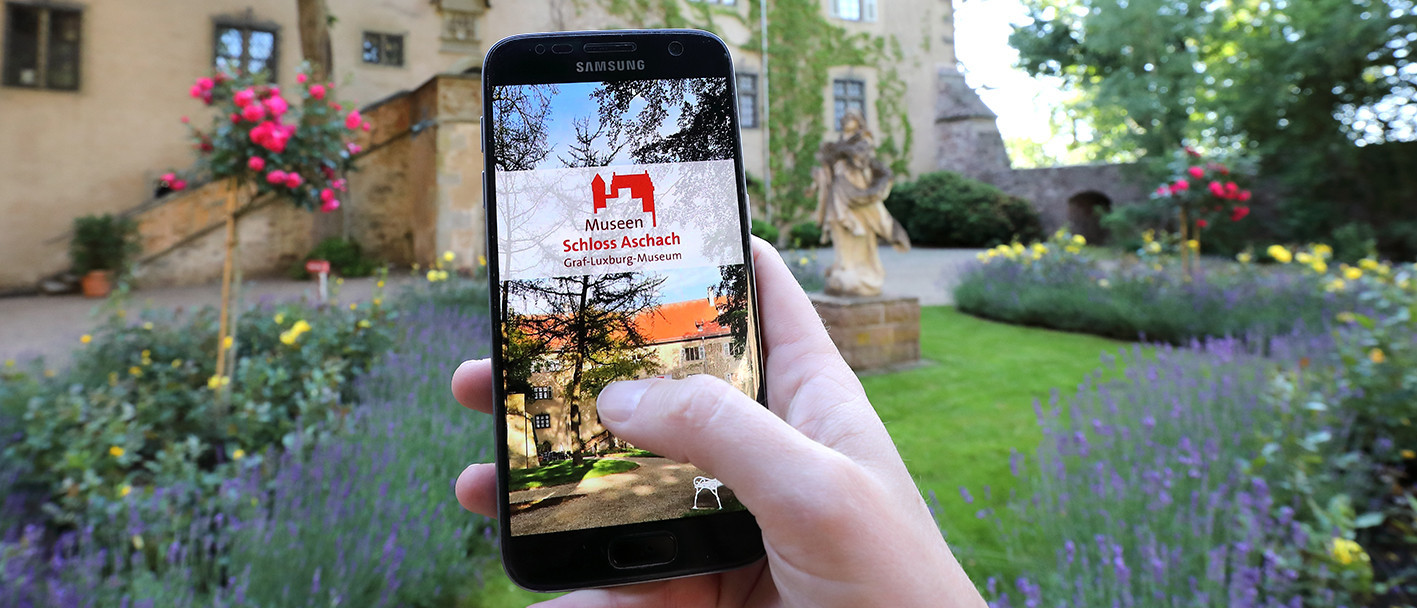 Eine Hand hält im Aschacher Schlosspark ein Smartphone. Auf dem Smartphone ist die Startseite der App &quot;Graf-Luxburg-Museum&quot; zu sehen.