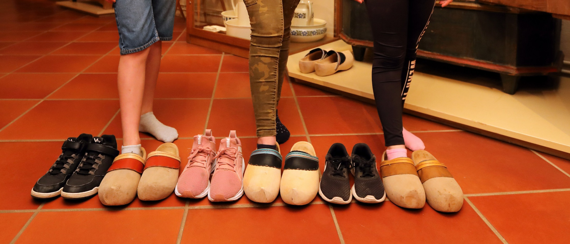Auf einem Boden stehen 6 Schuhpaare. Drei Paare sind moderne Turnschuhe und drei Paare Holzpantoffeln. Dahinter sieht man die Beine von drei Kindern.