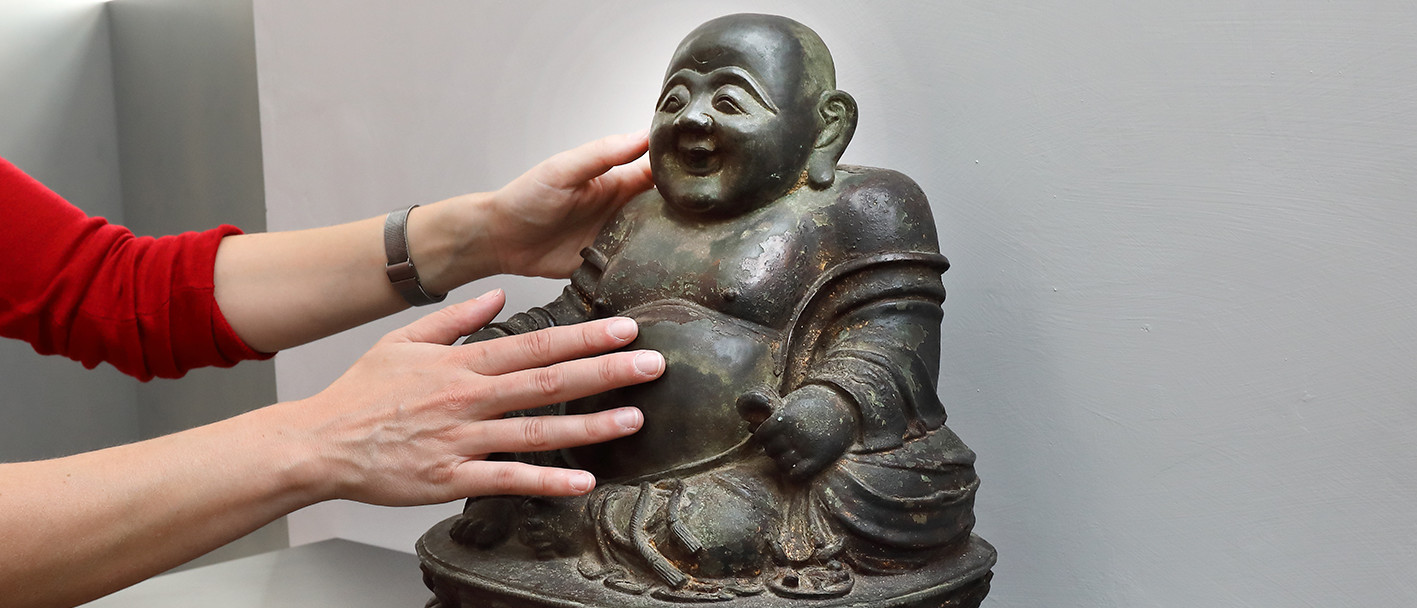 Zwei Hände kommen von links und berühren eine Budai-Skulptur aus Bronze.