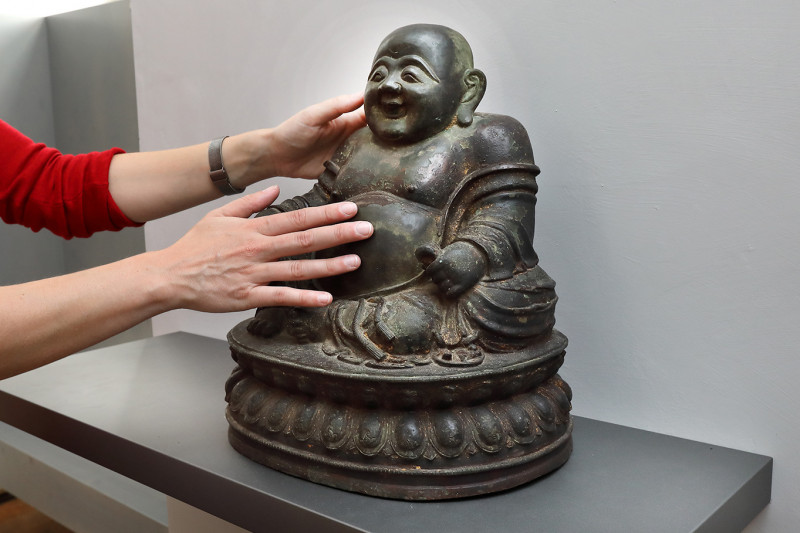 Zwei Hände kommen von links und berühren eine Budai-Skulptur aus Bronze.