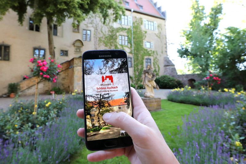 Eine Hand hält im Aschacher Schlosspark ein Smartphone. Auf dem Smartphone ist die Startseite der App &quot;Graf-Luxburg-Museum&quot; zu sehen.