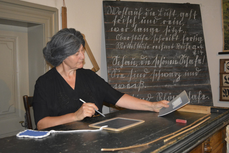 Eine Museumspädagogin hat sich als Schulfräulein verkleidet. Sie trägt ein schwarzes Kleid und eine Perücke. Sie sitzt am Lehrerpult und schaut ein Schulheft an. Im Hintergrund steht eine beschriebene Schultafel.
