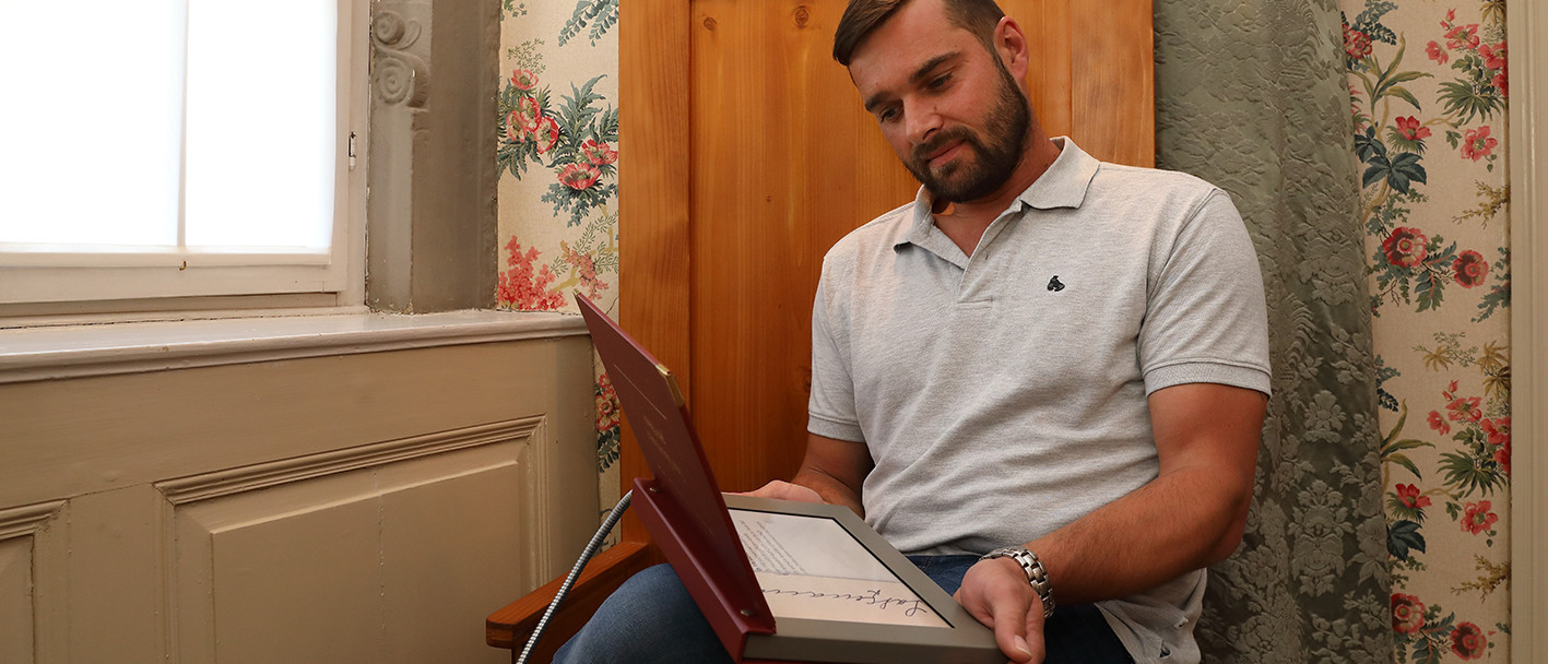 Ein Mann sitzt auf einer braunen Bank mit hoher Rückenlehne und hält ein Tablet in der Hand. Im Hintergrund ist die geblümte Tapete des Schlosssalons zu sehen.