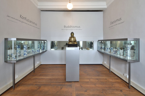 Das Foto zeigt einen Raum in der Ausstellung. In der Mitte steht eine Säule mit einer Figur aus Metall. Die Figur ist ein Buddha. Rund um den Buddha sind viele Sachen aus Porzellan ausgestellt. Das Porzellan ist bemalt.