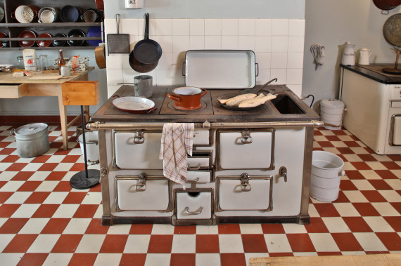 Auf dem Foto ist ein alter Herd zum Kochen. Der Herd wurde mit Holz beheizt. Auf dem Herd stehen Geschirr und Fische in einer Pfanne. Der Boden in der Küche hat rote und weiße Fliesen. Dahinter stehen ein Küchentisch und ein Wandregal.