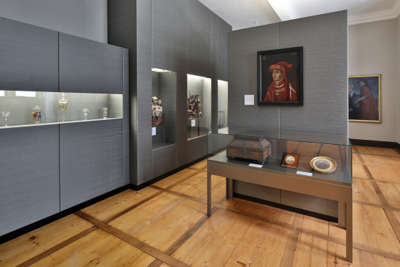 Das Foto zeigt einen Raum in der Ausstellung. Manche Wände und Regale sind grau. Figuren und Glasvasen sind hinter Glasscheiben zu sehen. Es gibt auch einige Gemälde.
