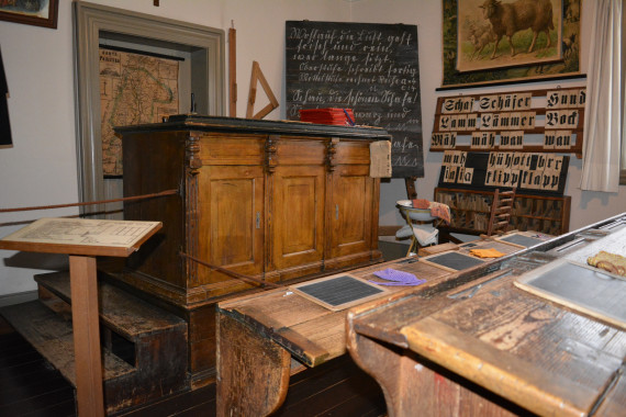 Auf dem Foto sind alte Schulbänke. Vorne ist ein großes Lehrerpult.  Neben dem Pult steht eine Tafel mit alter Schrift. Alle Möbel sind aus Holz. Das alte Schulzimmer sieht aus, als wären die Schulkinder gerade in der Pause.