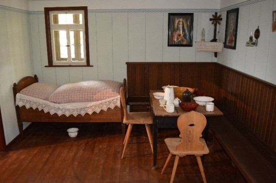 In einer kleinen Stube steht ein Bett unter einem Fenster. Unter dem Bett ist ein Nachttopf. Rechts sind eine Eckbank, ein Tisch und Stühle. Auf dem Tisch steht Essen. An der Wand sind ein Kreuz und Bilder.