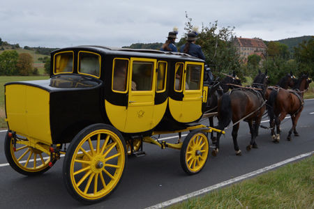 Foto von einer großen, alten Postkutsche. Die Kutsche ist gelb und schwarz bemalt. Vier Pferde ziehen die Kutsche auf einer Straße.