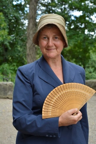 Auf dem Foto ist eine Dame mit einem Hut. Die Dame trägt ein blaues Kostüm und einen Fächer aus Holz. Die Dame sieht aus wie Gräfin Carola in der Vergangenheit.