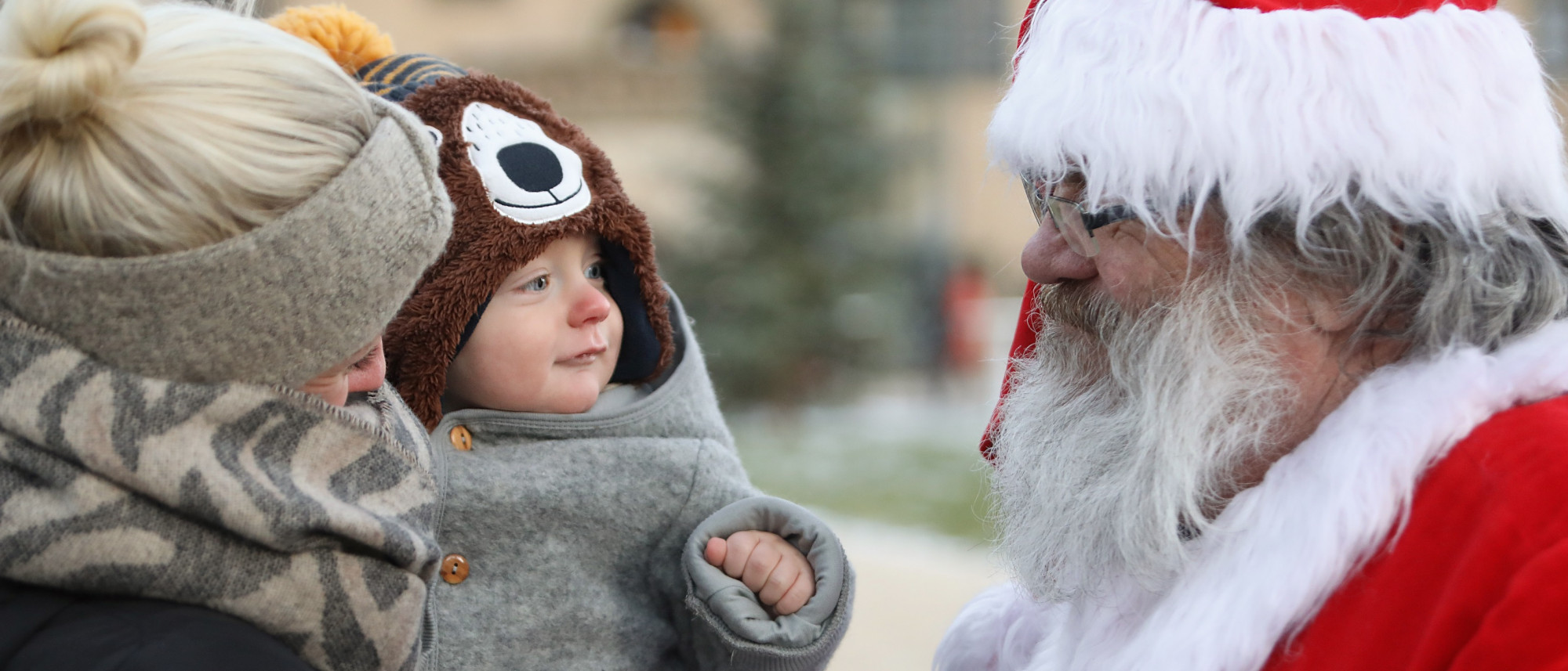 Ein kleines Kind auf dem Arm seiner Mama bekommt vom Weihnachtsmann eine Süßigkeit überreicht.
