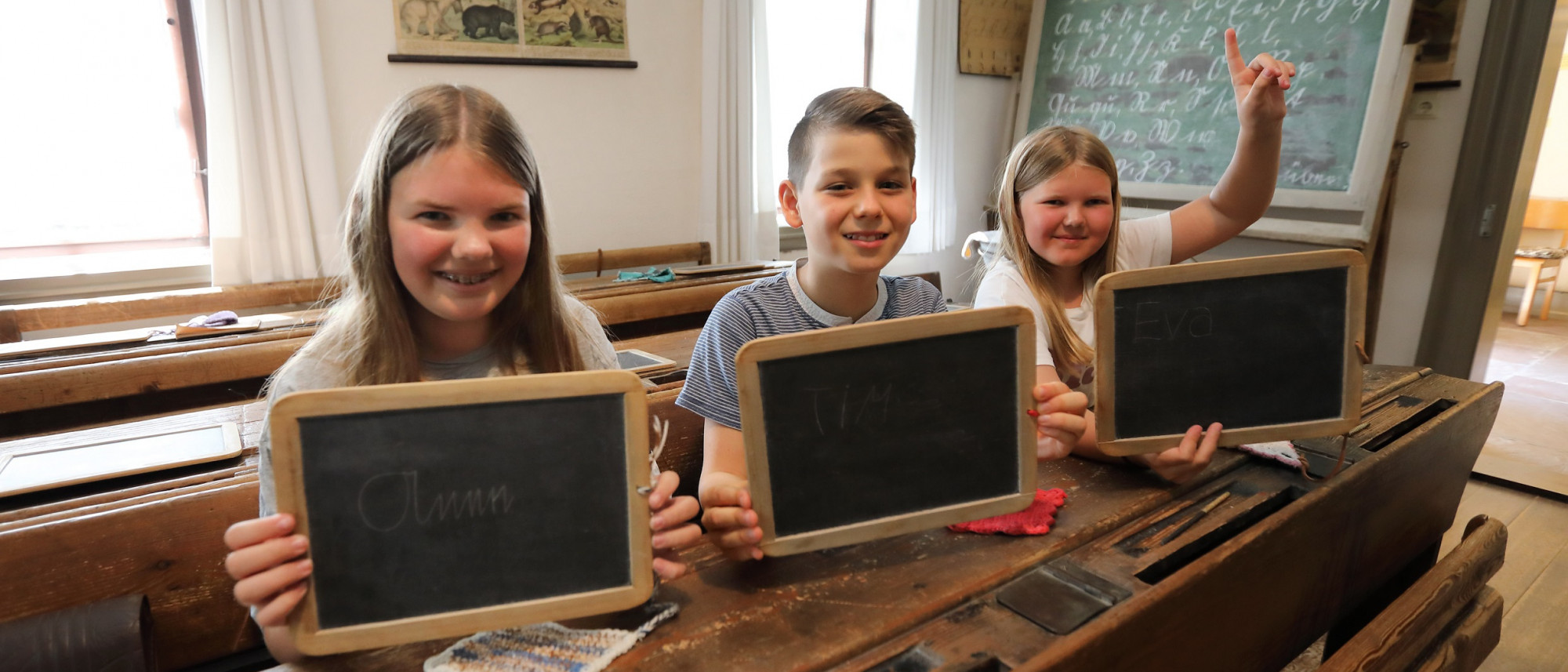 Drei Kinder sitzen auf einer Schulbank in einem alten Klassenzimmer. Sie halten jeweils eine Schiefertafel hoch, auf die sie mit Griffel ihre Namen geschrieben haben. Das Mädchen rechts außen meldet sich außerdem.