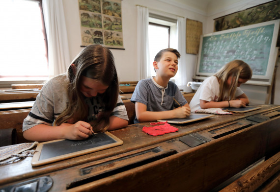3 Kinder sitzen auf einer alten Holzschulbank mit schräger Fläche. Sie schreiben mit Griffeln auf Schiefertafeln.