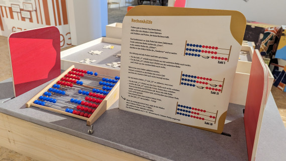 Links liegt ein Abakus, ein Rechenhilfsmittel mit roten und blauen Perlen. Daneben steht eine Texttafel, die den Abakus und seine Verwendung erklärt.