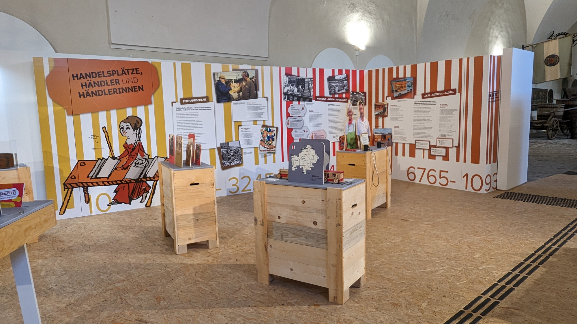 Ein Ausstellungsbereich mit farbigen Wänden und Ausstellungstexten und Bildern sowie Holzsockeln, auf denen Texttafeln und Mitmachstationen sind.